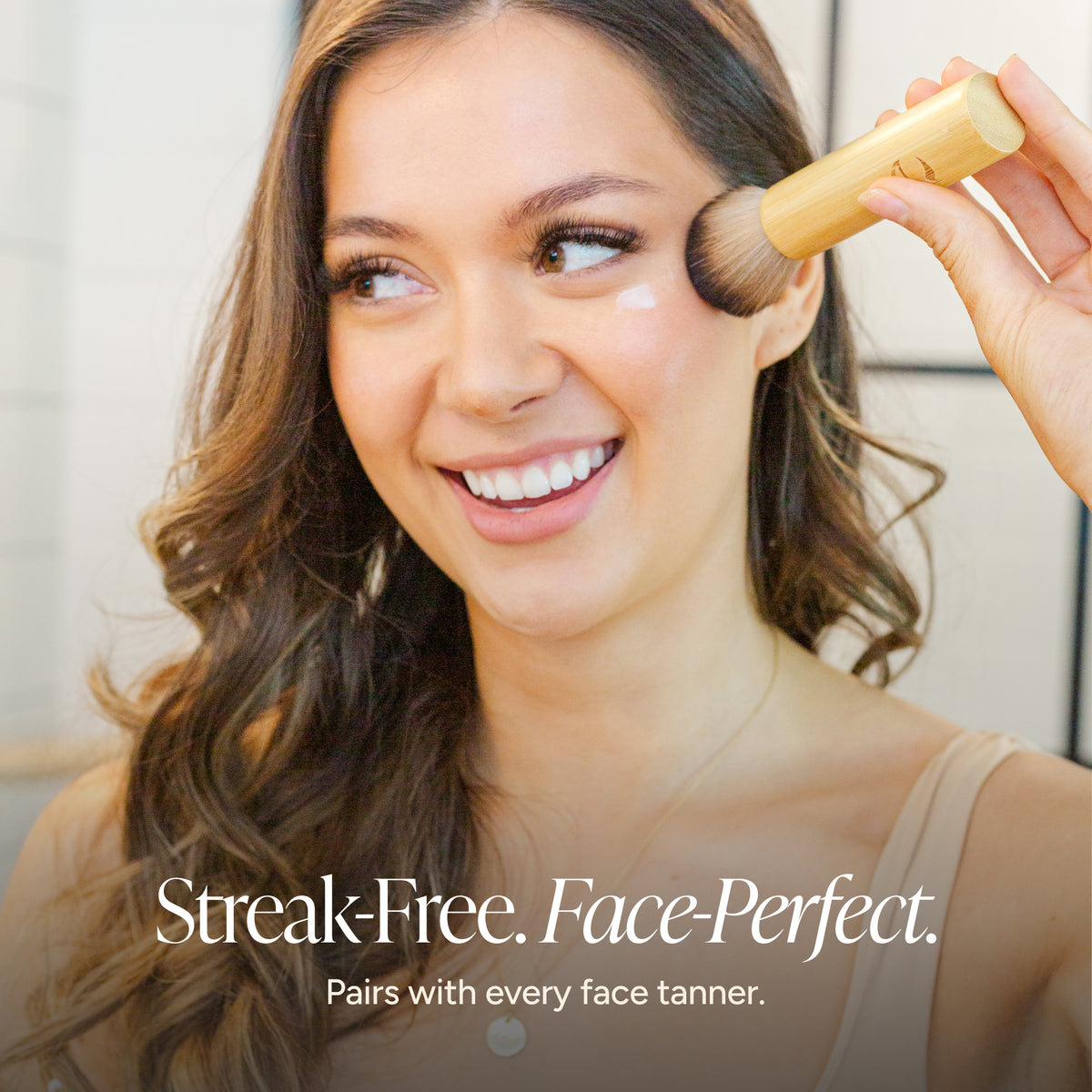 A smiling woman with long brown hair applies face tan using a kabuki brush with a bambii handle. Text at the bottom reads 'Streak-Free.