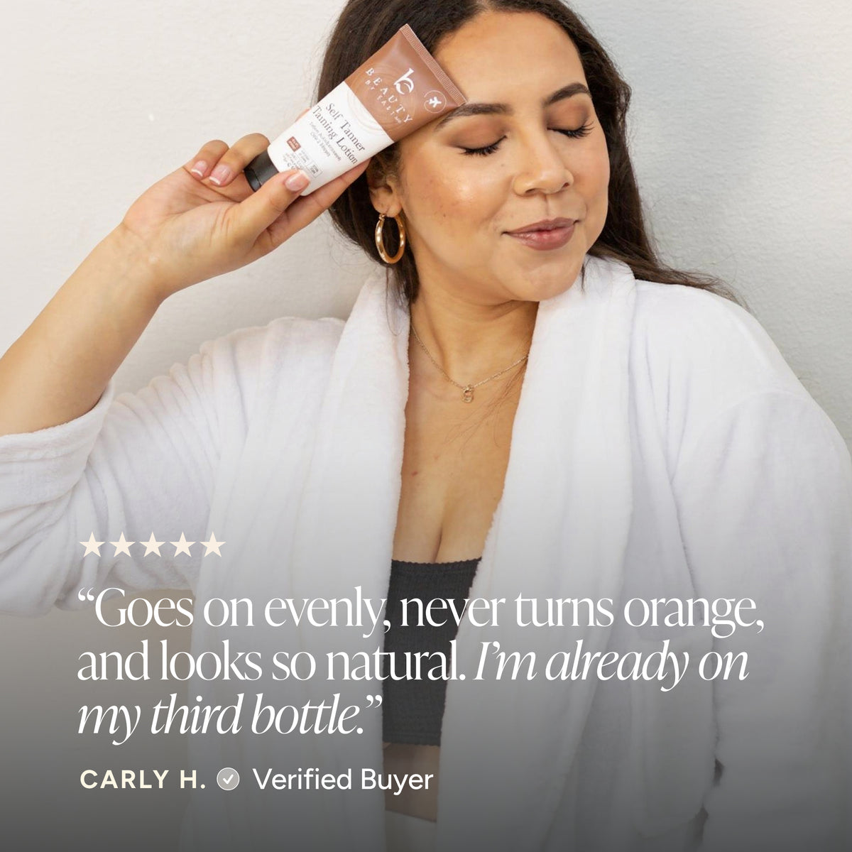 A woman in a white robe holds a tube of self tanner tanning lotion near her face.