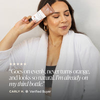 A woman in a white robe holds a tube of self tanner tanning lotion near her face.