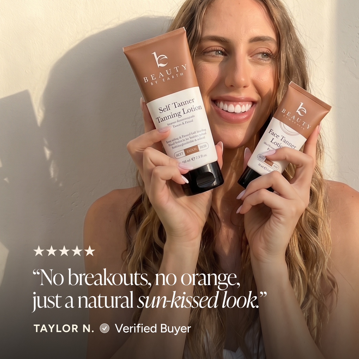 A smiling woman holds two tubes of Beauty by Earth Self Tanning Tanning Lotion against a warm sunlit wall.