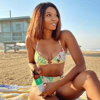 Woman in a bikini sitting on the beach with a bottle of sunscreen.