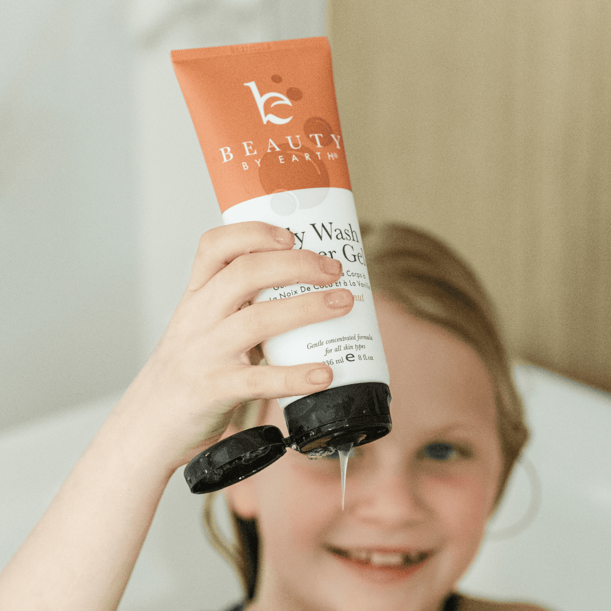 Child holding a tube of 'Beauty by Earth' body wash shower gel in vanilla coconut with visible brand name.