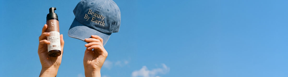 Hand holding a bottle of skincare product and a cap with 'Beauty By Earth' text against a clear blue sky.
