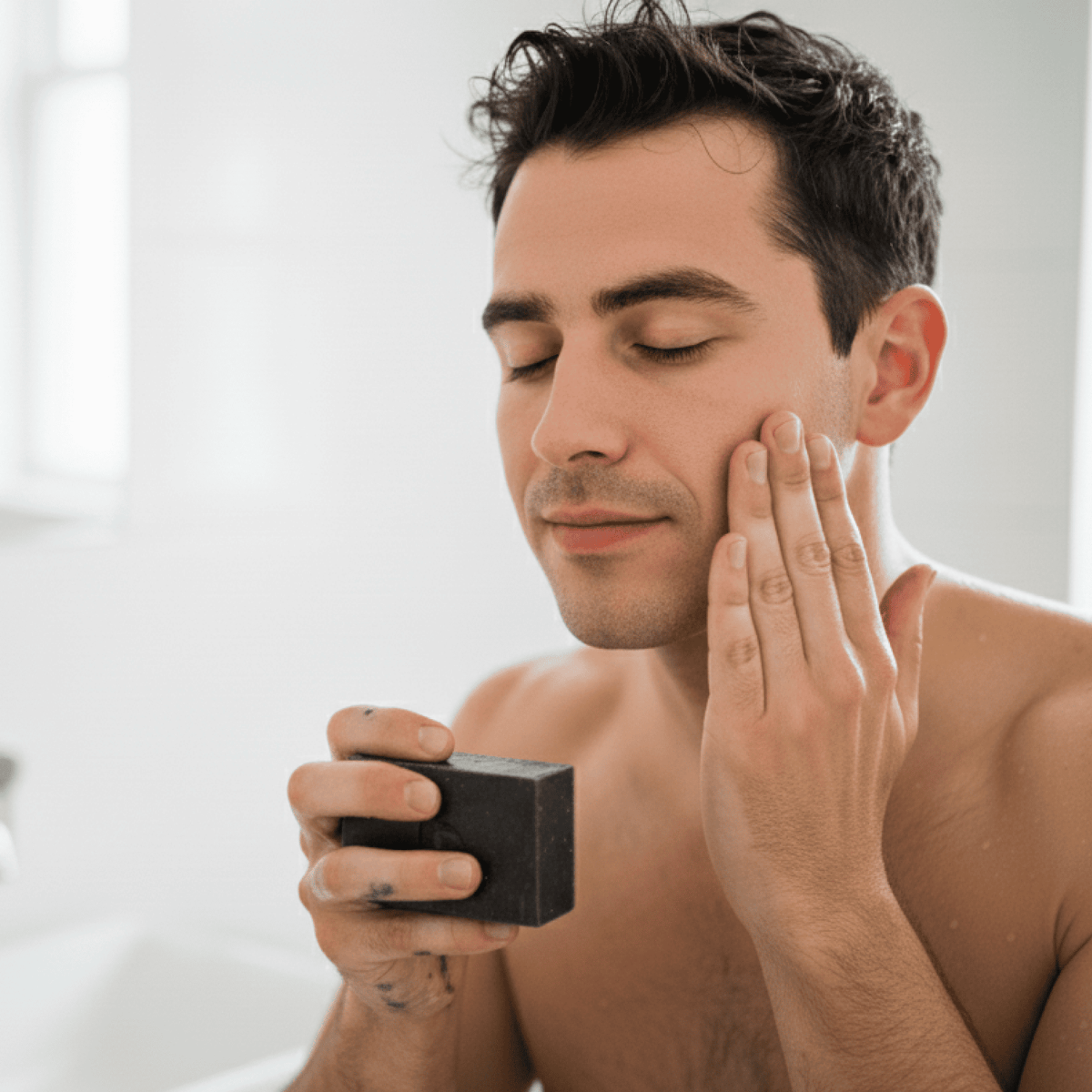 Man applying charcoal soap to his face with a bar of charcoal soap in hand in a bathroom setting