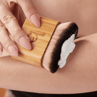 Person using a wooden brush with bristles on a person's arm, applying a white cream or lotion.