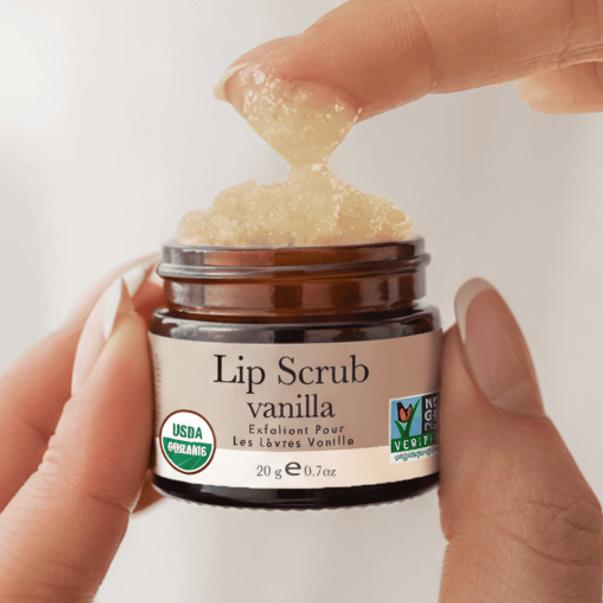 Lip scrub jar with vanilla flavor held by a hand against a neutral background