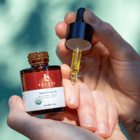 Hand holding a bottle of Beauty by Earth nail and cuticle oil with a dropper filled with yellow liquid.