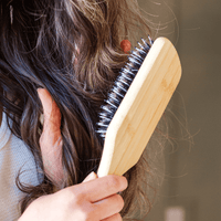Person using a wooden hairbrush with bristles to detangle hair against a neutral background