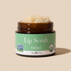 An open amber glass jar of lip scrub with a green label reading 'Lip Scrub mint' on a warm beige background. The scrub is visible at the top of the jar, and certification logos for Non-GMO Project and USDA Organic appear on the label.