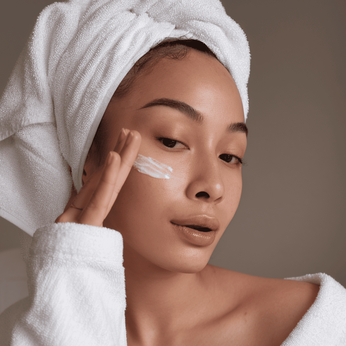 Woman applying cream to her face with a towel on her head against a neutral background